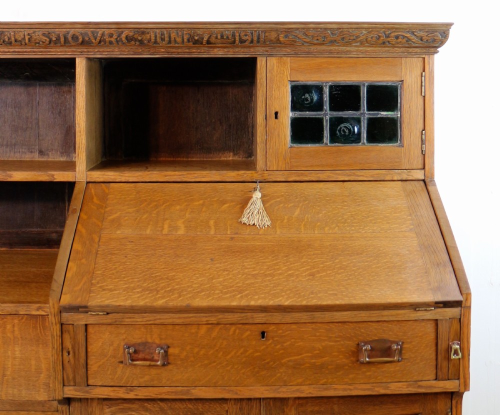 An Arts and Crafts quarter-sawn oak bureau bookcase featuring an asymmetrical design with open shelves, a fall-front writing slope, a leaded green glass cabinet, and drawers with copper handles. View VII