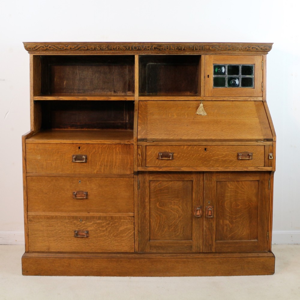 An Arts and Crafts quarter-sawn oak bureau bookcase featuring an asymmetrical design with open shelves, a fall-front writing slope, a leaded green glass cabinet, and drawers with copper handles. View XII