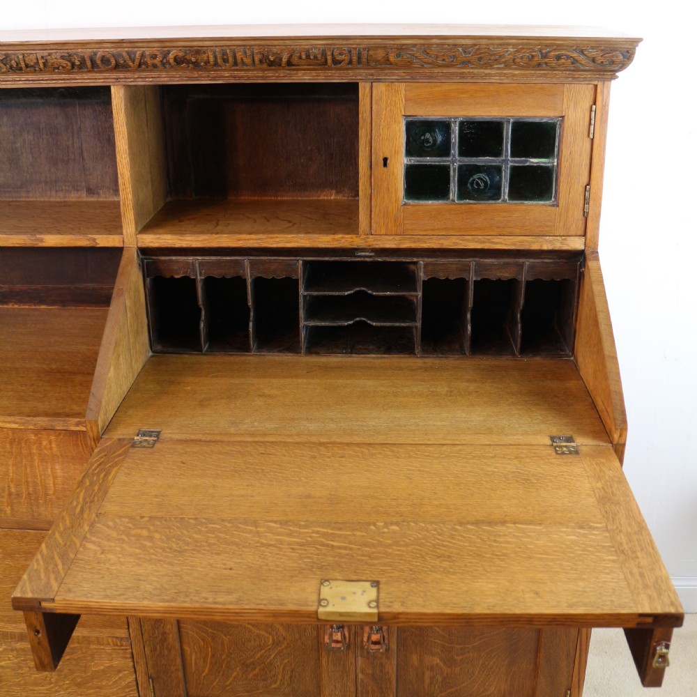 An Arts and Crafts quarter-sawn oak bureau bookcase featuring an asymmetrical design with open shelves, a fall-front writing slope, a leaded green glass cabinet, and drawers with copper handles. View XIV