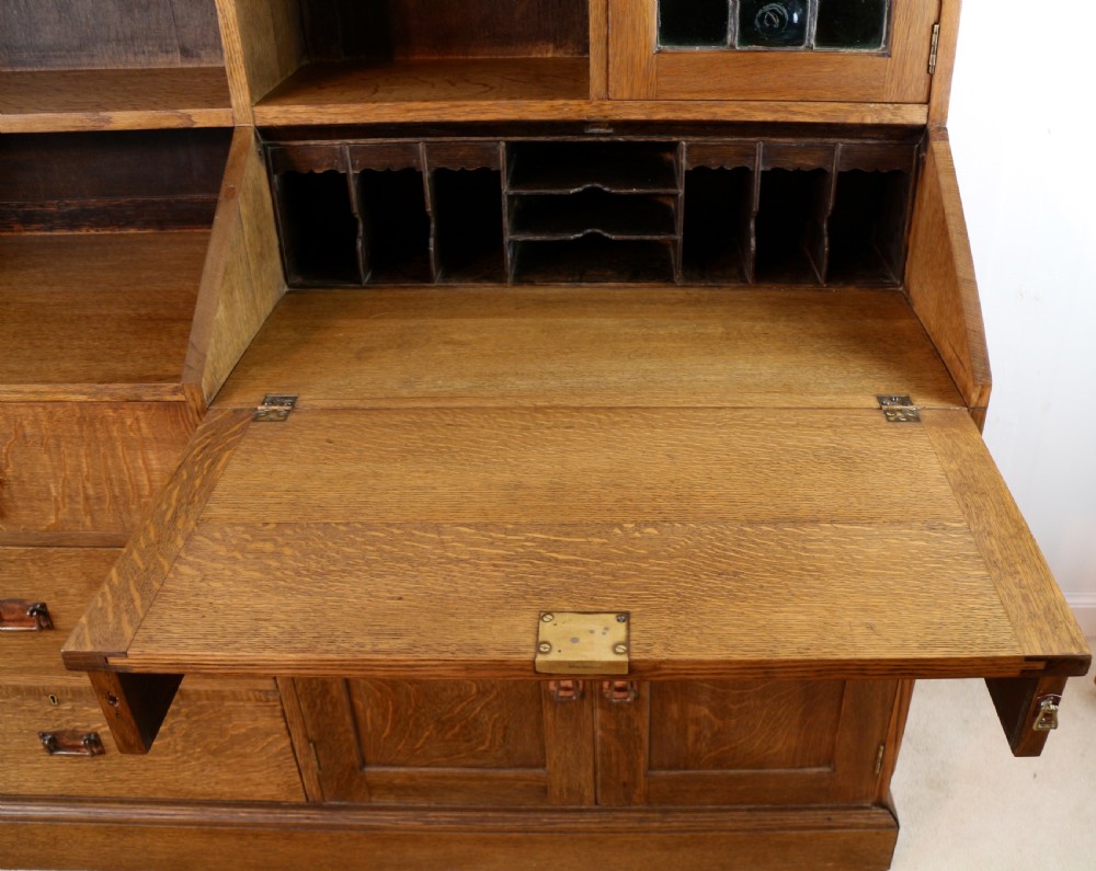 An Arts and Crafts quarter-sawn oak bureau bookcase featuring an asymmetrical design with open shelves, a fall-front writing slope, a leaded green glass cabinet, and drawers with copper handles. View XVI