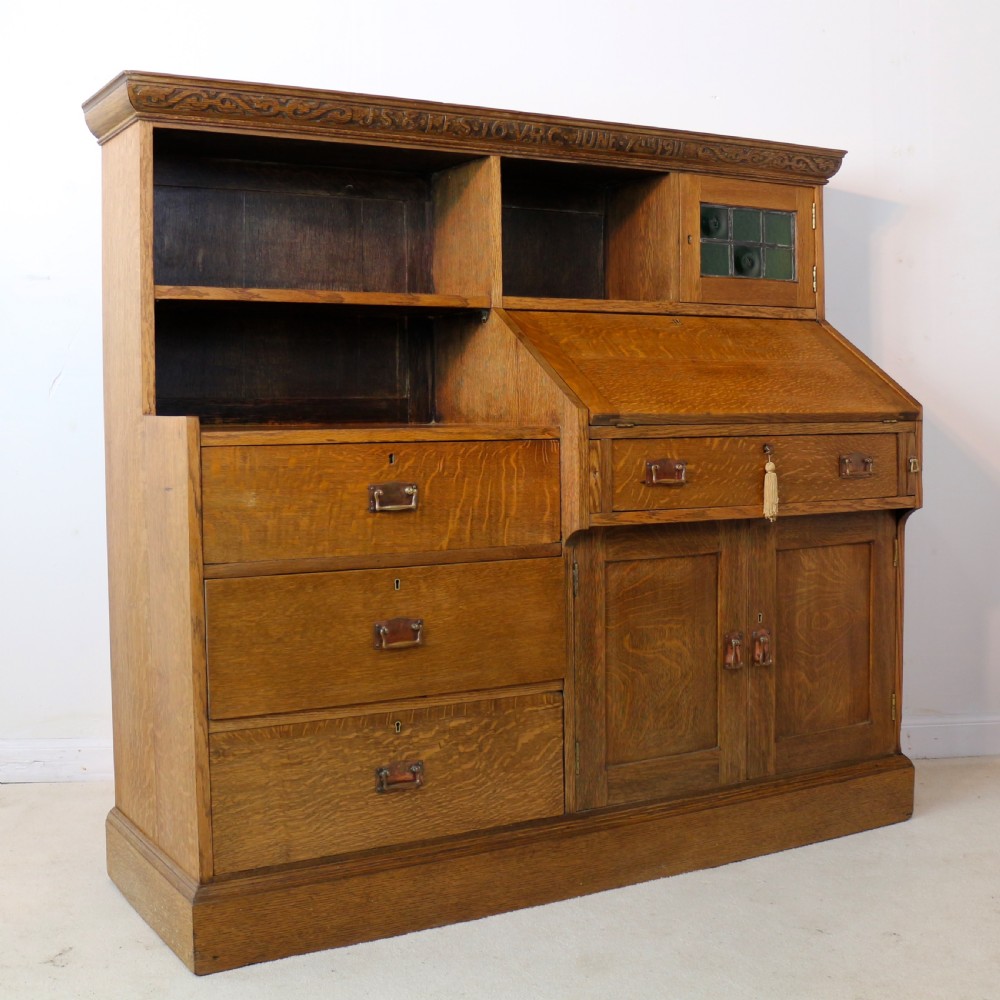 An Arts and Crafts quarter-sawn oak bureau bookcase featuring an asymmetrical design with open shelves, a fall-front writing slope, a leaded green glass cabinet, and drawers with copper handles. View XVII