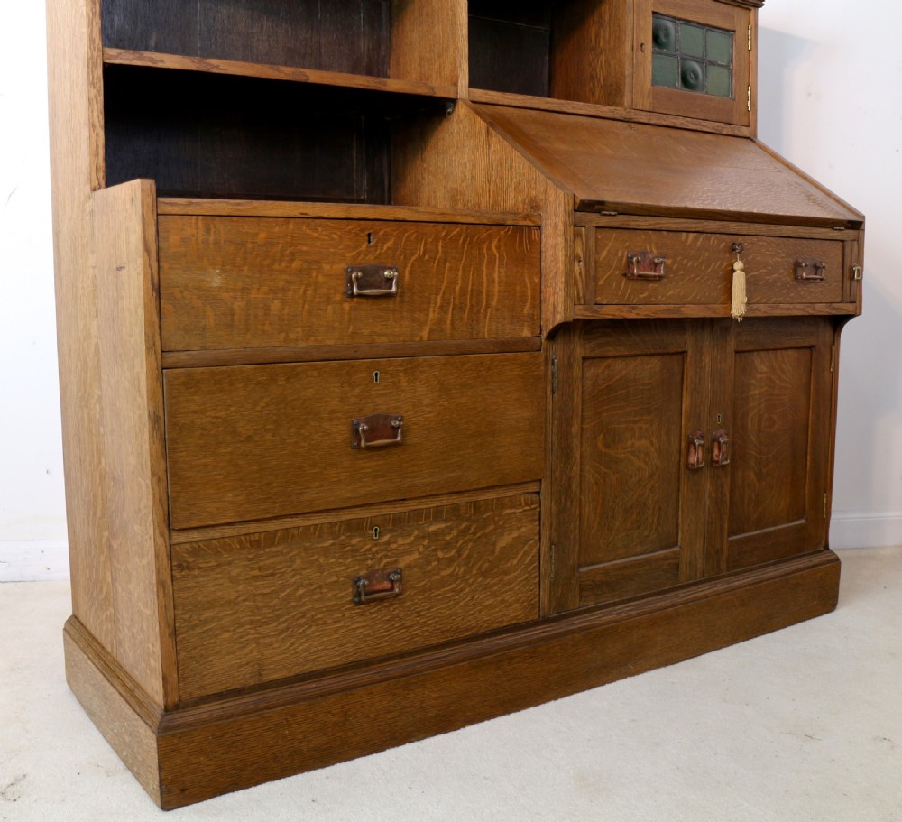An Arts and Crafts quarter-sawn oak bureau bookcase featuring an asymmetrical design with open shelves, a fall-front writing slope, a leaded green glass cabinet, and drawers with copper handles. View XIX