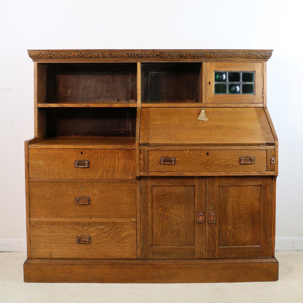 An Arts and Crafts quarter-sawn oak bureau bookcase featuring an asymmetrical design with open shelves, a fall-front writing slope, a leaded green glass cabinet, and drawers with copper handles. View XX
