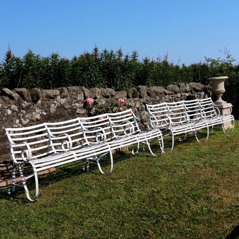 A pair of long white painted wrought iron Victorian garden benches with slatted backs and seats and scrolled armrests.