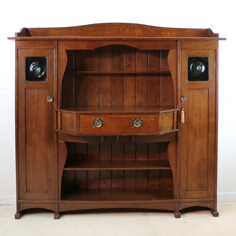 An Arts and Crafts solid oak sideboard featuring a central drawer, open shelving, and two side doors accented with green bullseye glass roundels.