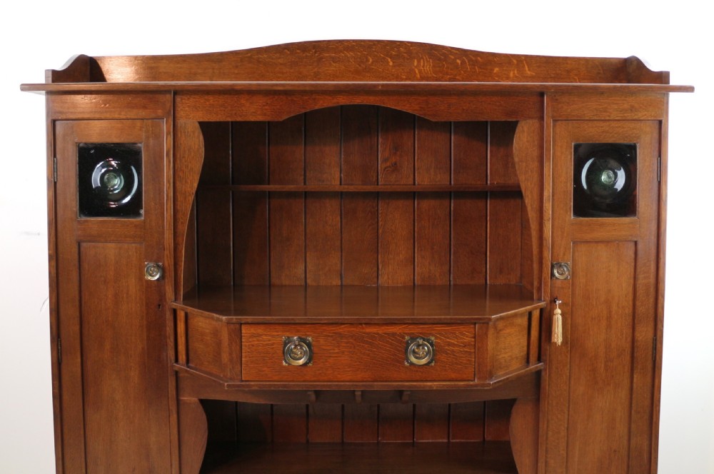 An Arts and Crafts solid oak sideboard featuring a central drawer, open shelving, and two side doors accented with green bullseye glass roundels. View II