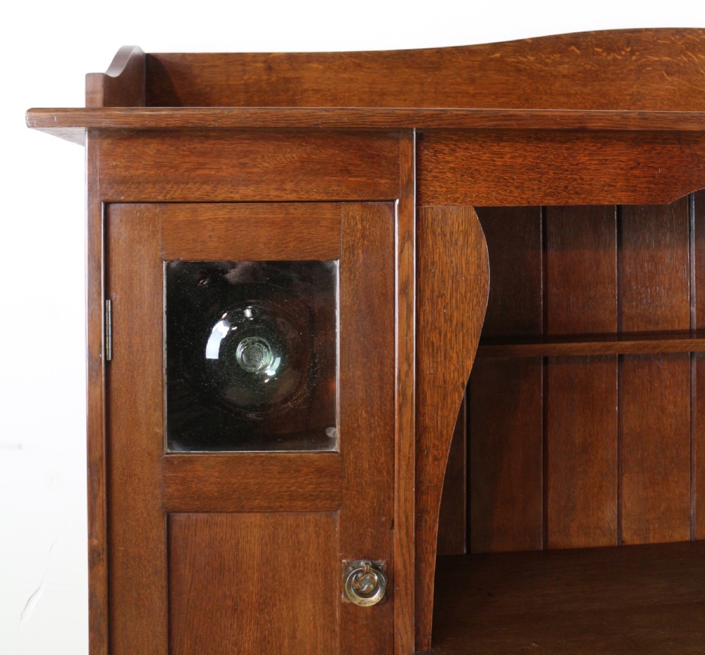 An Arts and Crafts solid oak sideboard featuring a central drawer, open shelving, and two side doors accented with green bullseye glass roundels. View III