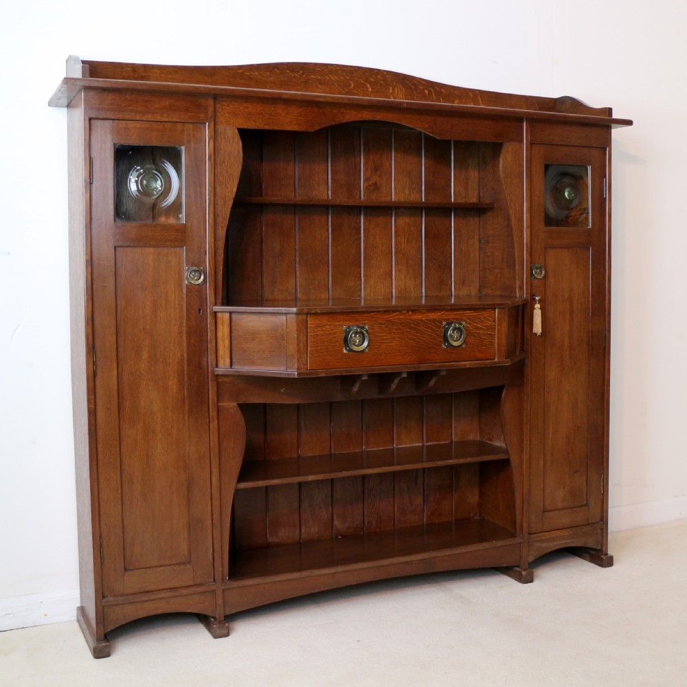 An Arts and Crafts solid oak sideboard featuring a central drawer, open shelving, and two side doors accented with green bullseye glass roundels. View VI