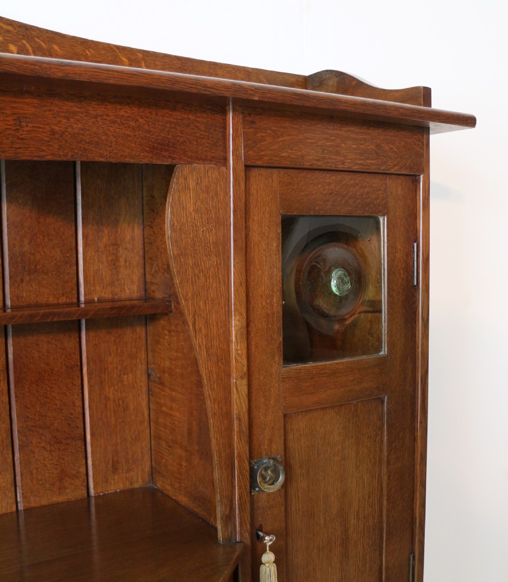 An Arts and Crafts solid oak sideboard featuring a central drawer, open shelving, and two side doors accented with green bullseye glass roundels. View IX