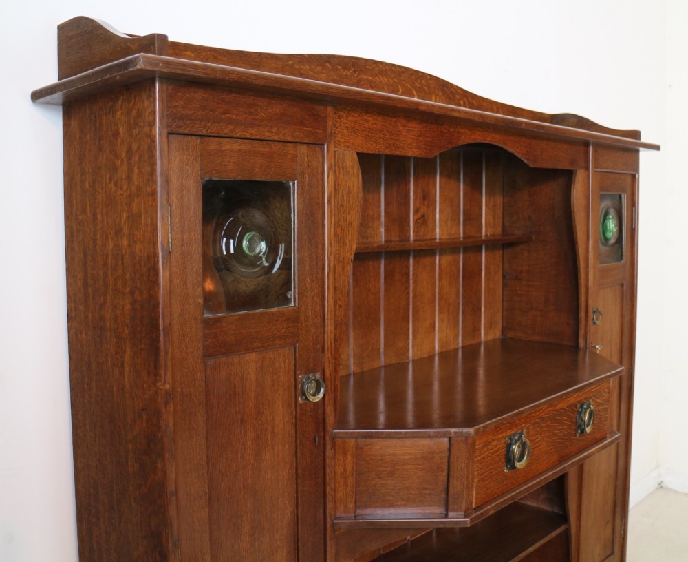 An Arts and Crafts solid oak sideboard featuring a central drawer, open shelving, and two side doors accented with green bullseye glass roundels. View X