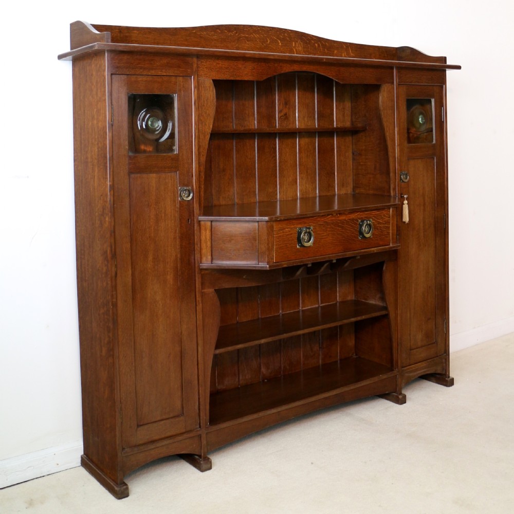 An Arts and Crafts solid oak sideboard featuring a central drawer, open shelving, and two side doors accented with green bullseye glass roundels. View XI