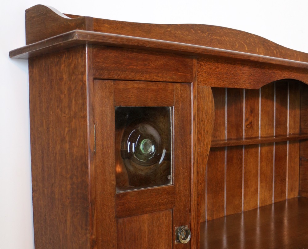 An Arts and Crafts solid oak sideboard featuring a central drawer, open shelving, and two side doors accented with green bullseye glass roundels. View XIII