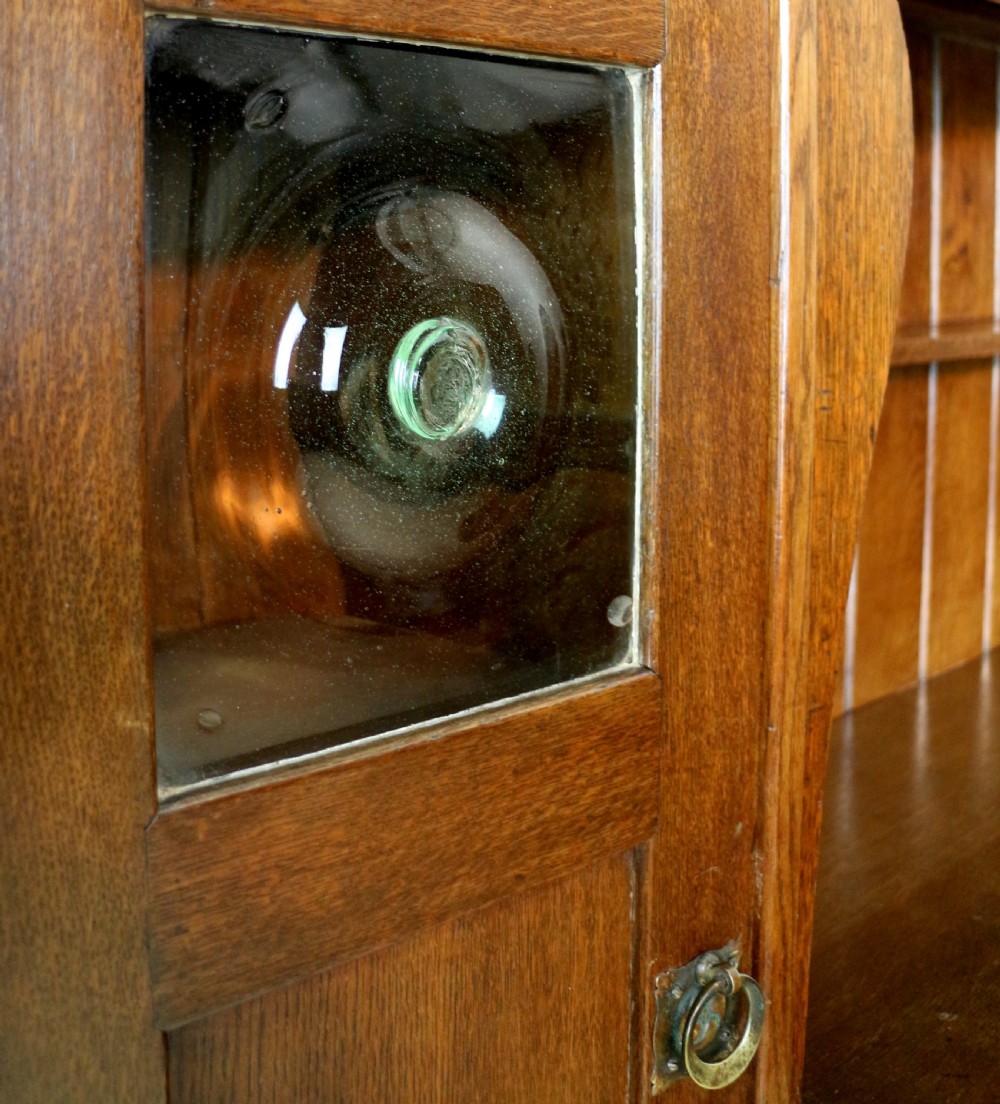 An Arts and Crafts solid oak sideboard featuring a central drawer, open shelving, and two side doors accented with green bullseye glass roundels. View XIV