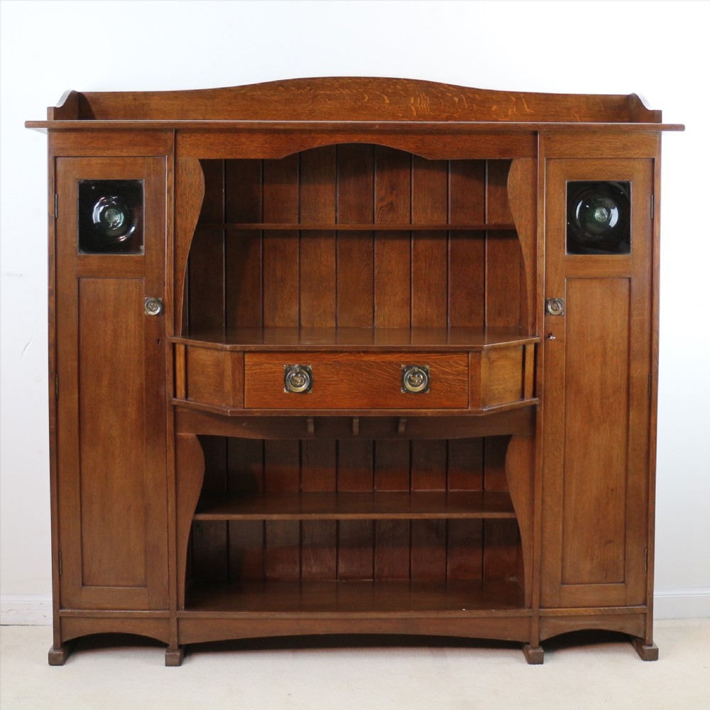 An Arts and Crafts solid oak sideboard featuring a central drawer, open shelving, and two side doors accented with green bullseye glass roundels. View XV