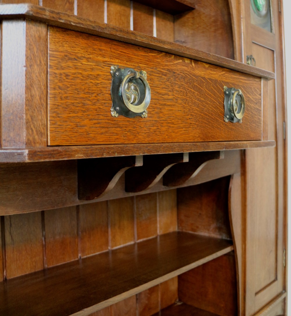 An Arts and Crafts solid oak sideboard featuring a central drawer, open shelving, and two side doors accented with green bullseye glass roundels. View XVI