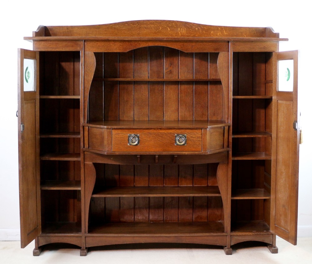 An Arts and Crafts solid oak sideboard featuring a central drawer, open shelving, and two side doors accented with green bullseye glass roundels. View XVII
