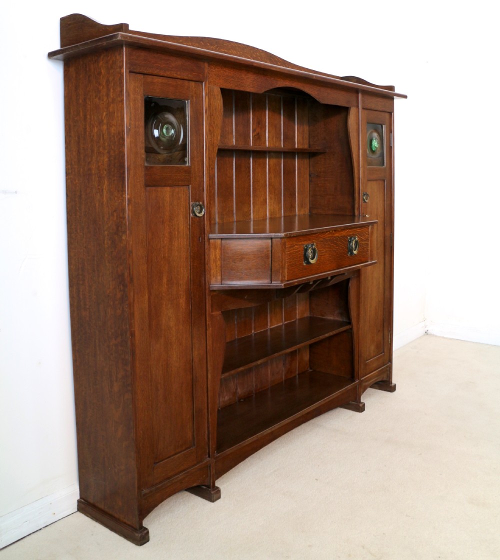 An Arts and Crafts solid oak sideboard featuring a central drawer, open shelving, and two side doors accented with green bullseye glass roundels. View XX