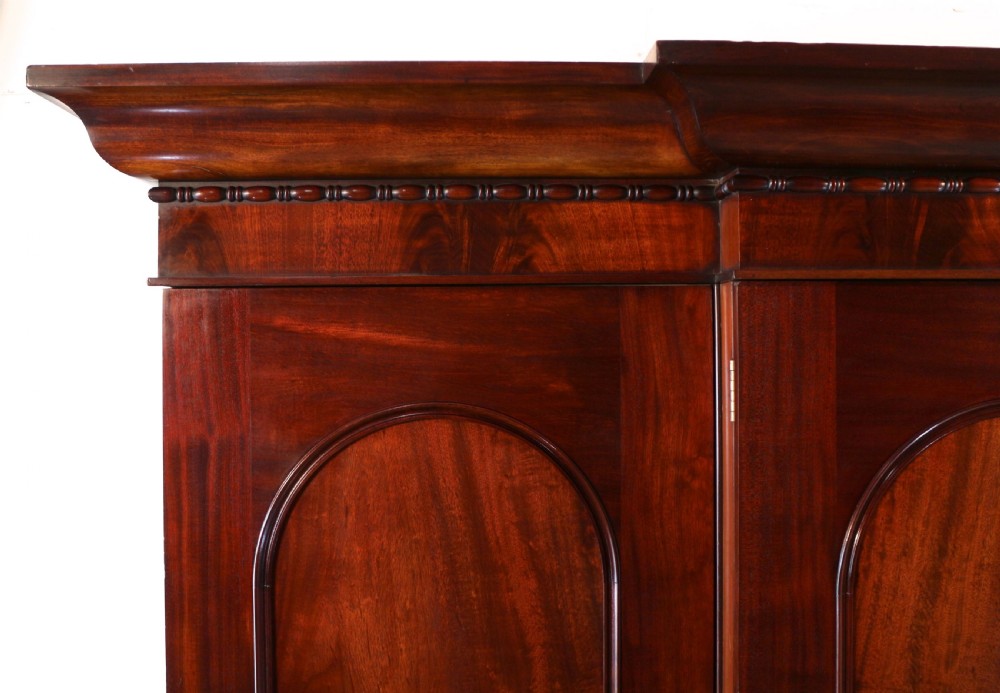A nineteenth-century breakfront wardrobe featuring flame mahogany veneer, four arched panel doors, and a moulded projecting cornice. View V