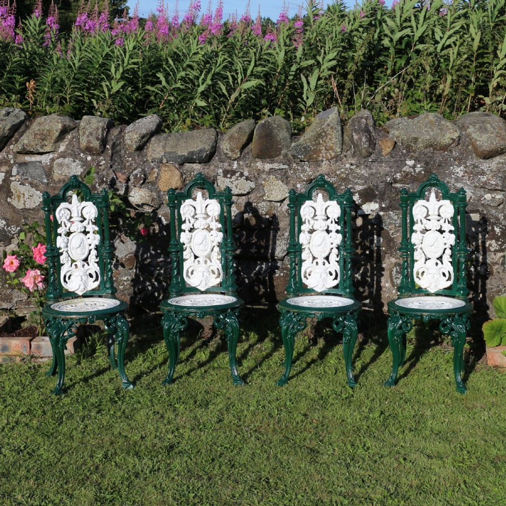 A set of four Victorian cast iron garden chairs featuring white openwork backrests with central medallion cameos framed by dark green structural frames and cabriole legs.