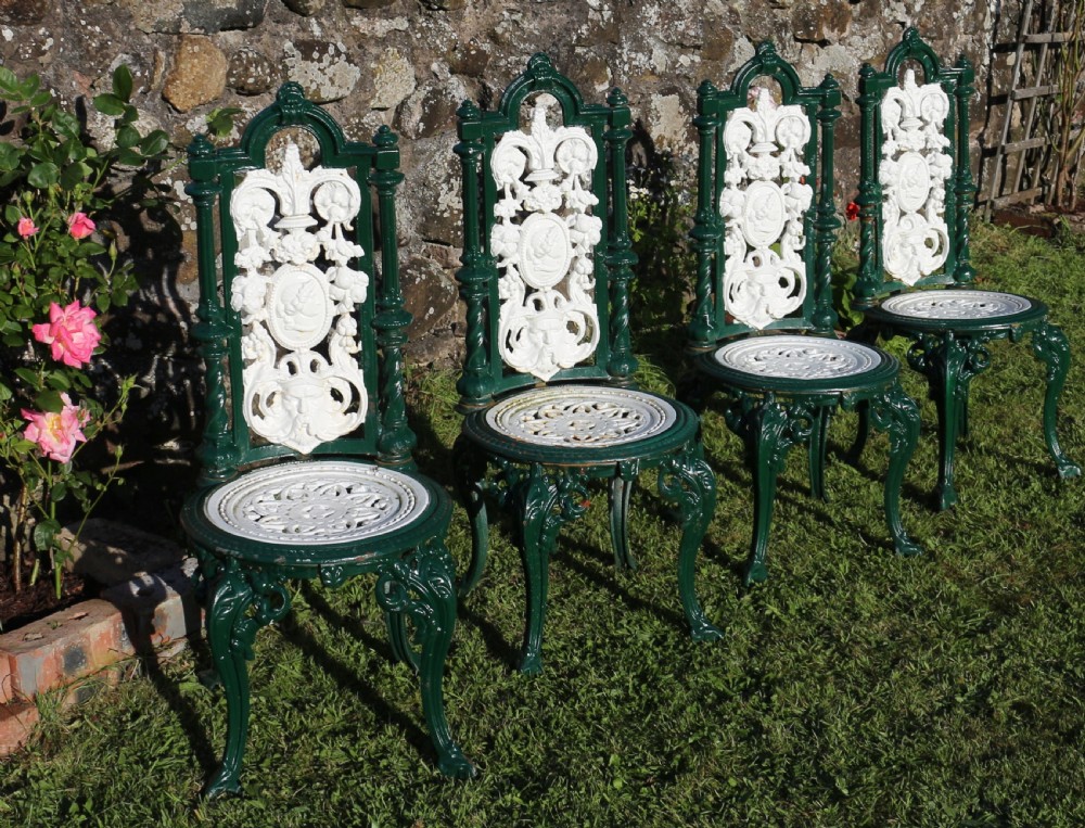 A set of four Victorian cast iron garden chairs featuring white openwork backrests with central medallion cameos framed by dark green structural frames and cabriole legs. View V