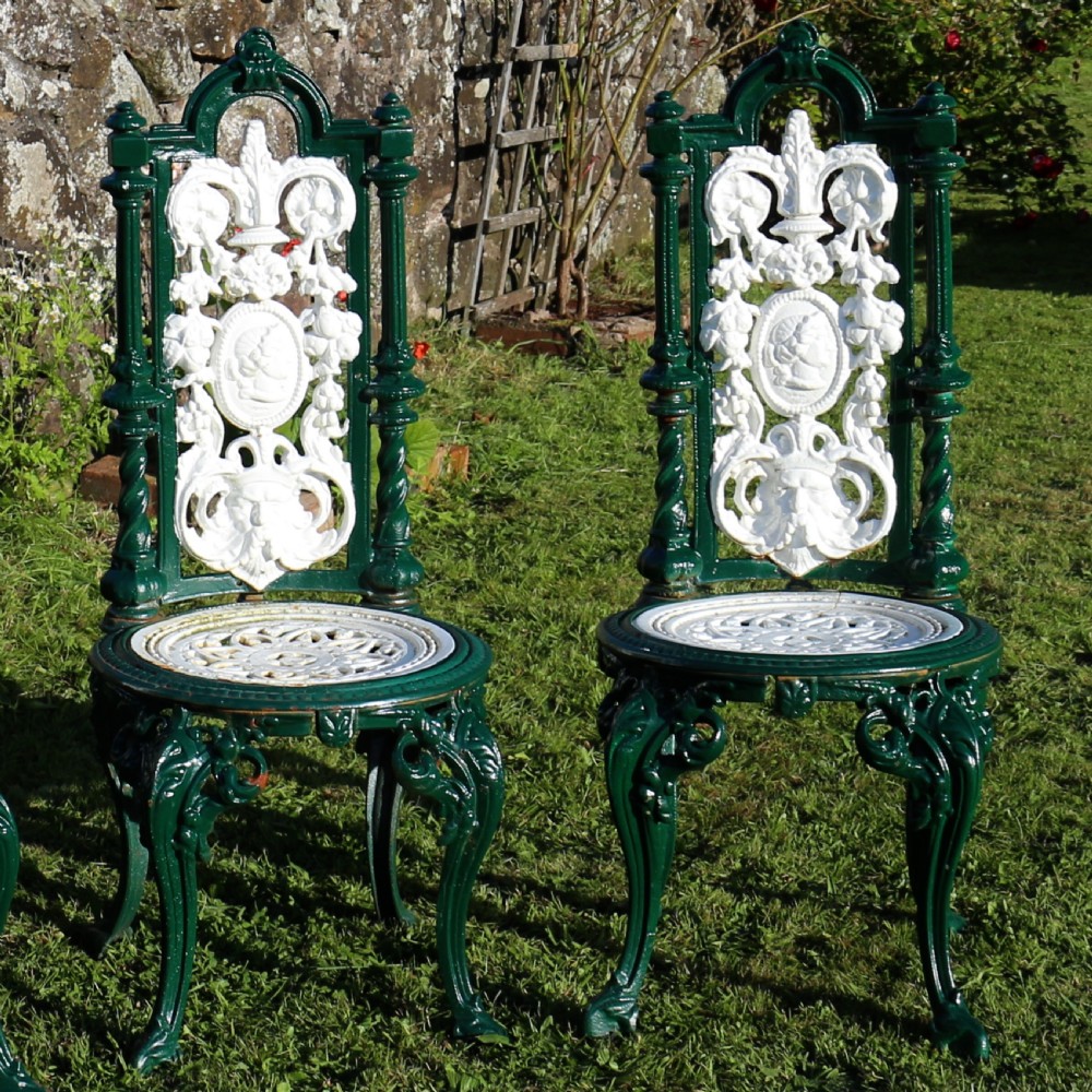 A set of four Victorian cast iron garden chairs featuring white openwork backrests with central medallion cameos framed by dark green structural frames and cabriole legs. View VII