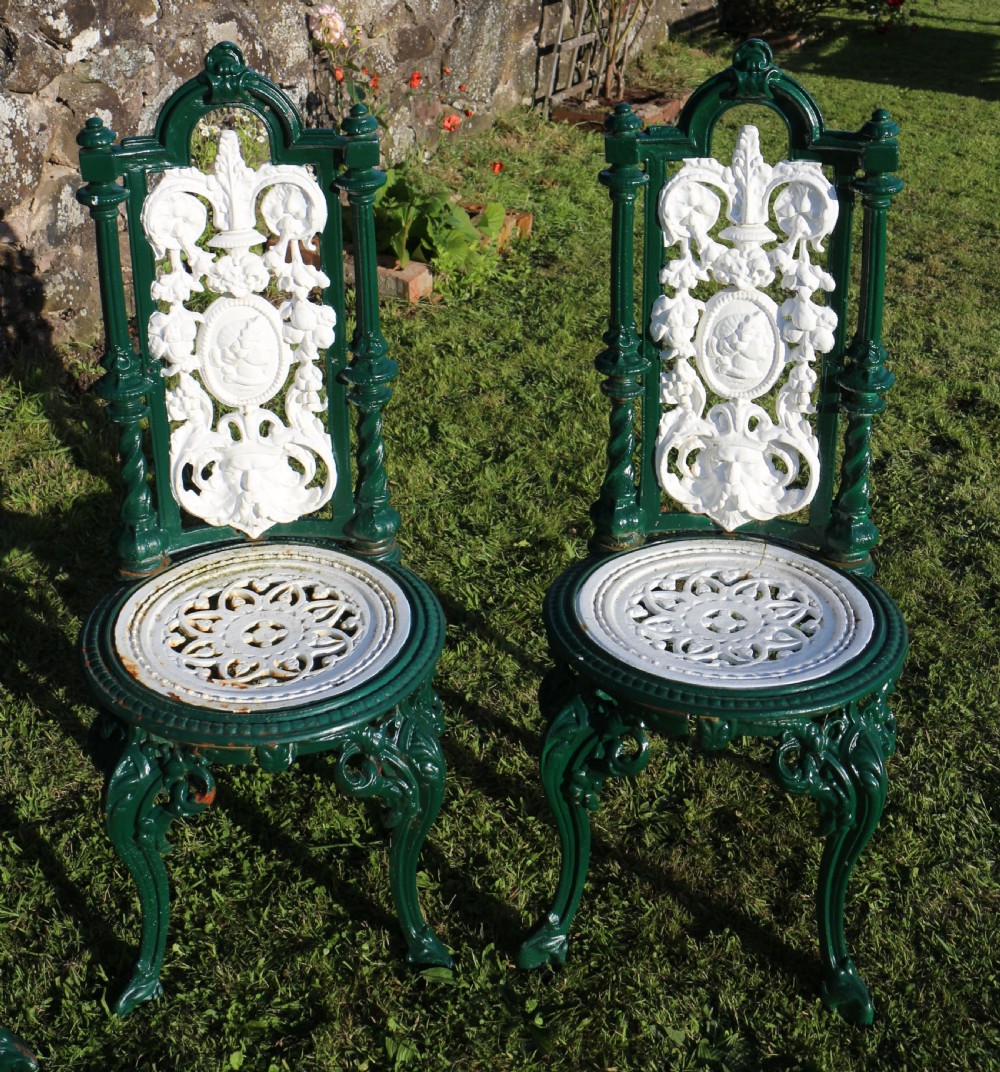 A set of four Victorian cast iron garden chairs featuring white openwork backrests with central medallion cameos framed by dark green structural frames and cabriole legs. View X