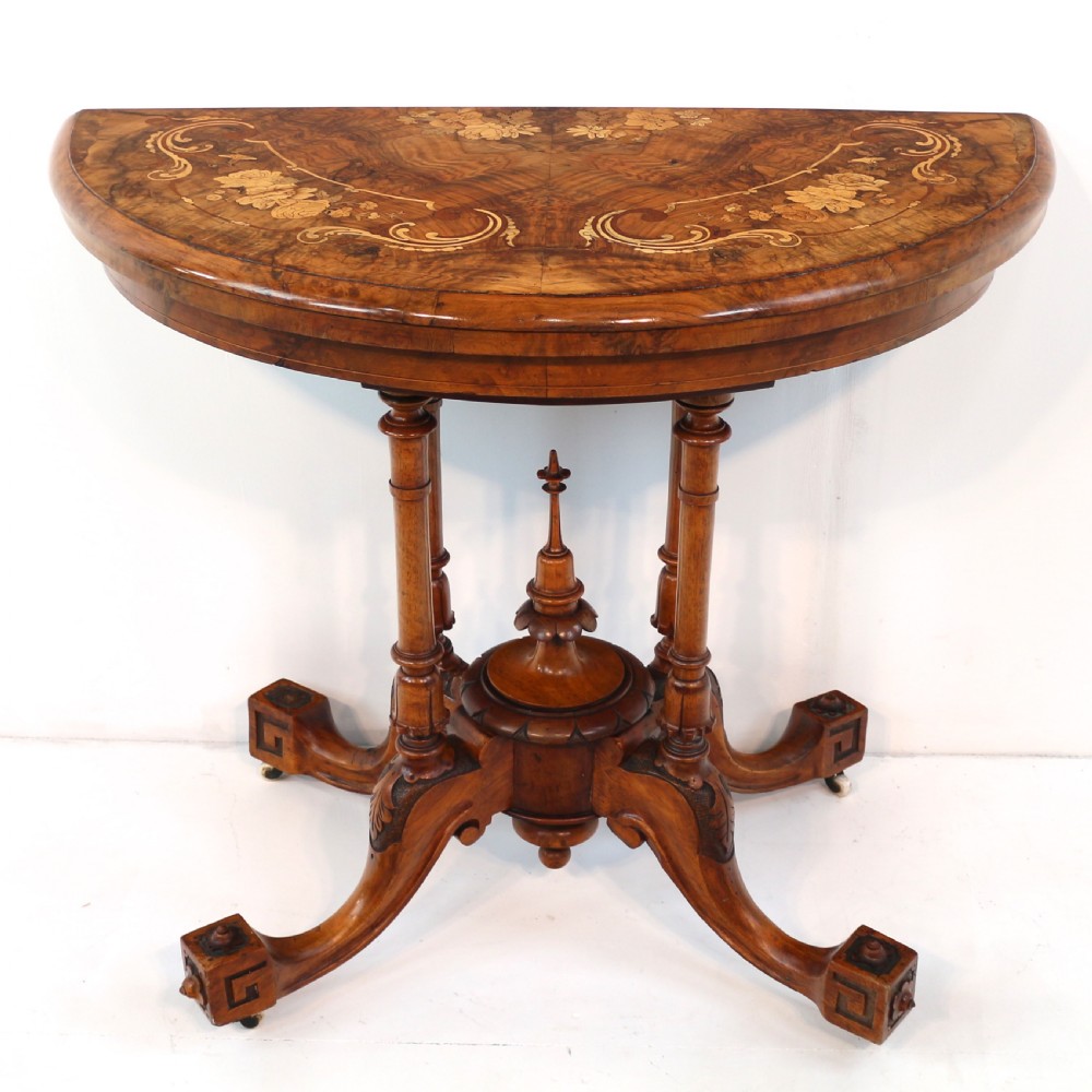 A Victorian burr walnut demilune card table featuring floral marquetry inlays, turned twin-column supports, and a carved central finial on quadruped legs.