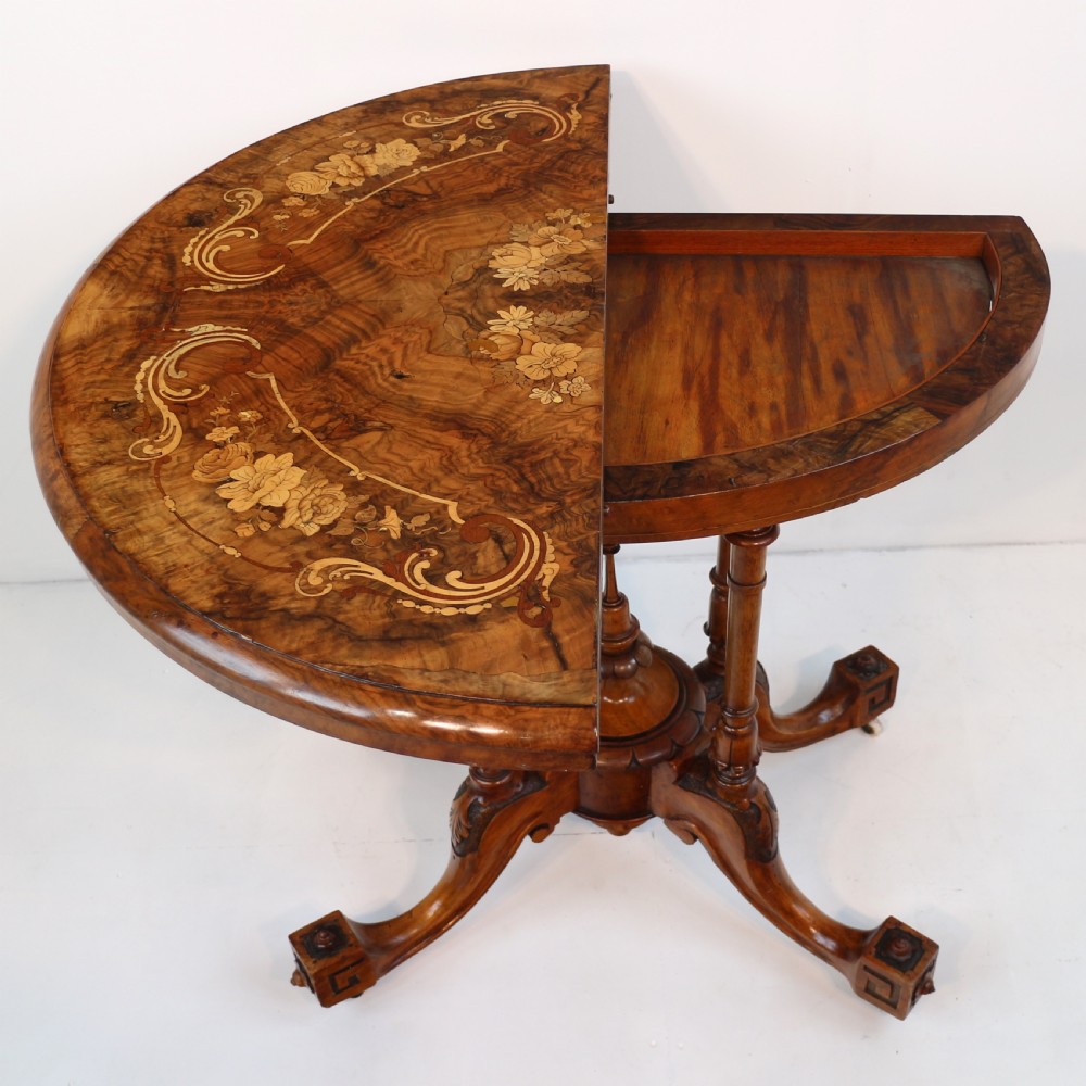 A Victorian burr walnut demilune card table featuring floral marquetry inlays, turned twin-column supports, and a carved central finial on quadruped legs. View V
