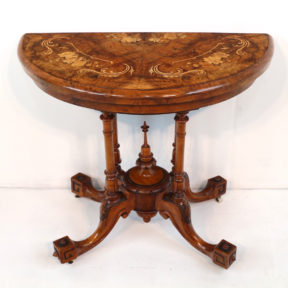 A Victorian burr walnut demilune card table featuring floral marquetry inlays, turned twin-column supports, and a carved central finial on quadruped legs. View X