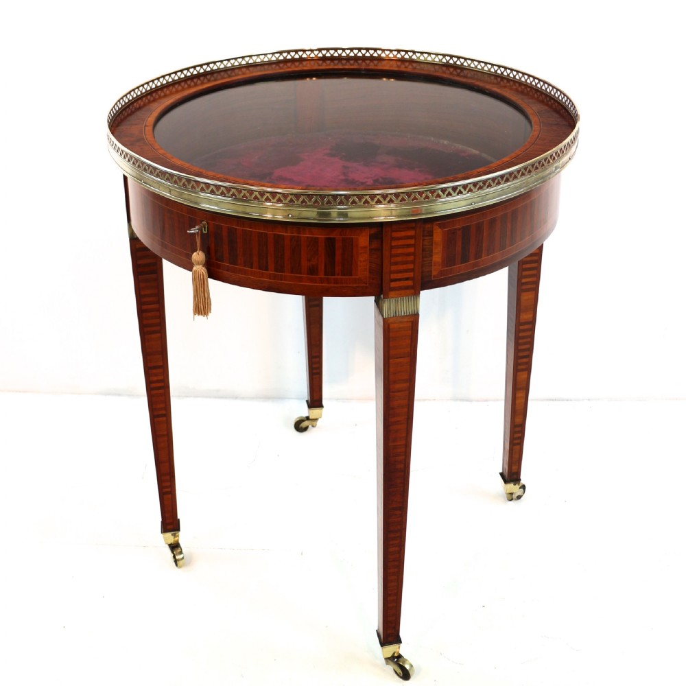 A circular Victorian rosewood bijouterie table featuring a glazed display lid, pierced brass rim gallery, inlaid tapered legs, and brass castors.