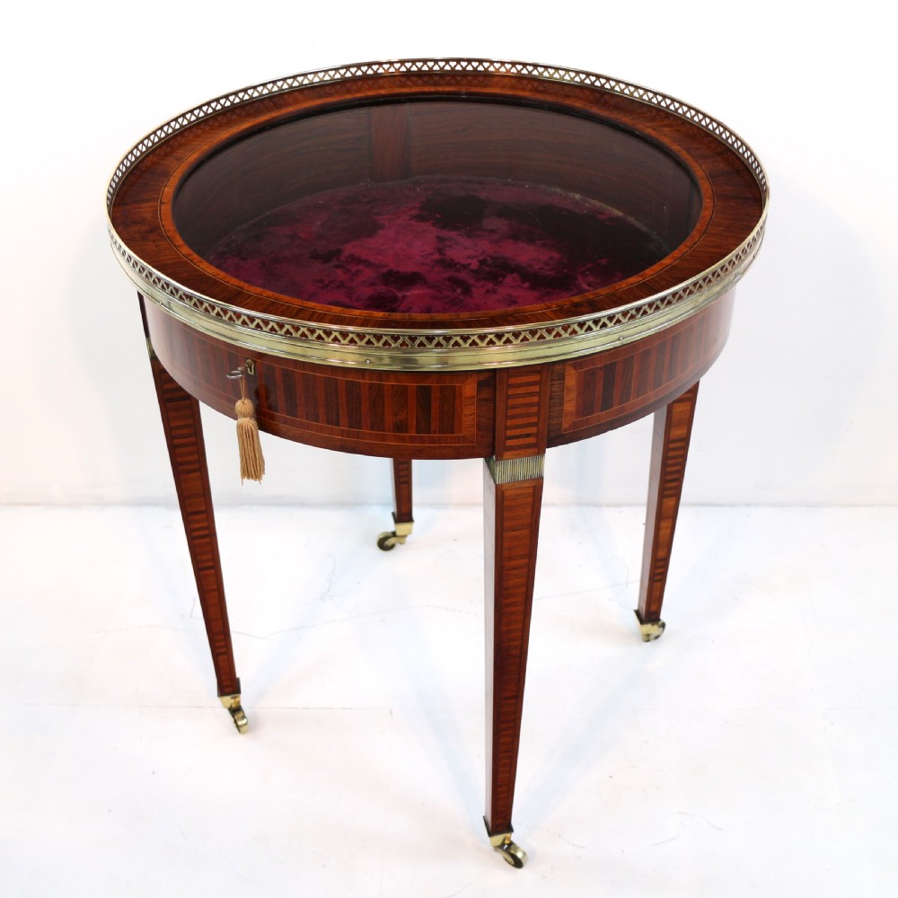 A circular Victorian rosewood bijouterie table featuring a glazed display lid, pierced brass rim gallery, inlaid tapered legs, and brass castors. View II