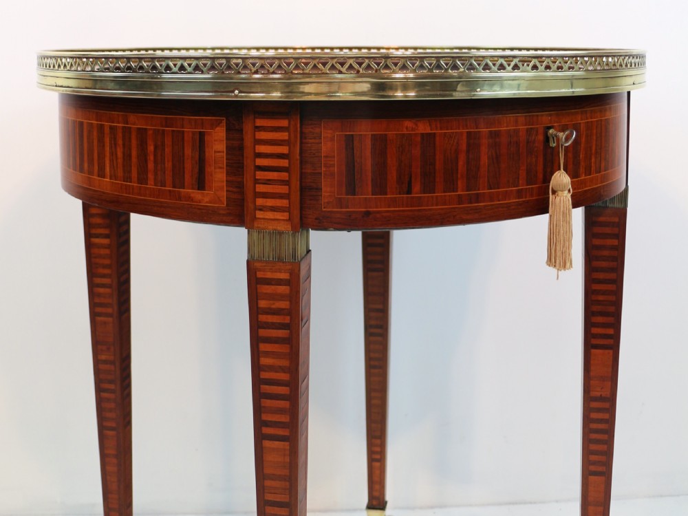 A circular Victorian rosewood bijouterie table featuring a glazed display lid, pierced brass rim gallery, inlaid tapered legs, and brass castors. View VI