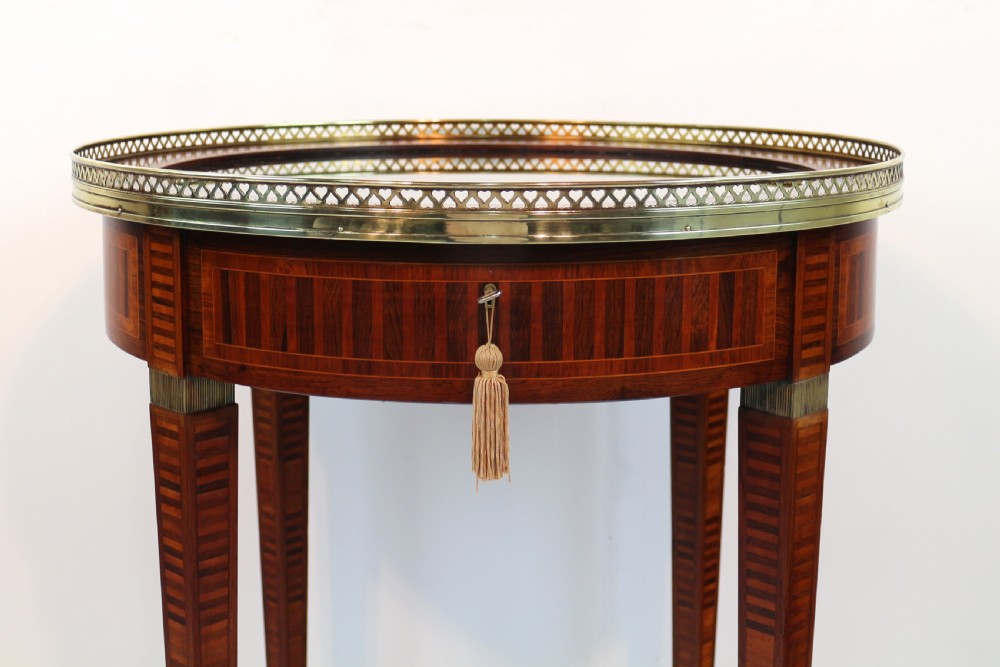 A circular Victorian rosewood bijouterie table featuring a glazed display lid, pierced brass rim gallery, inlaid tapered legs, and brass castors. View VII