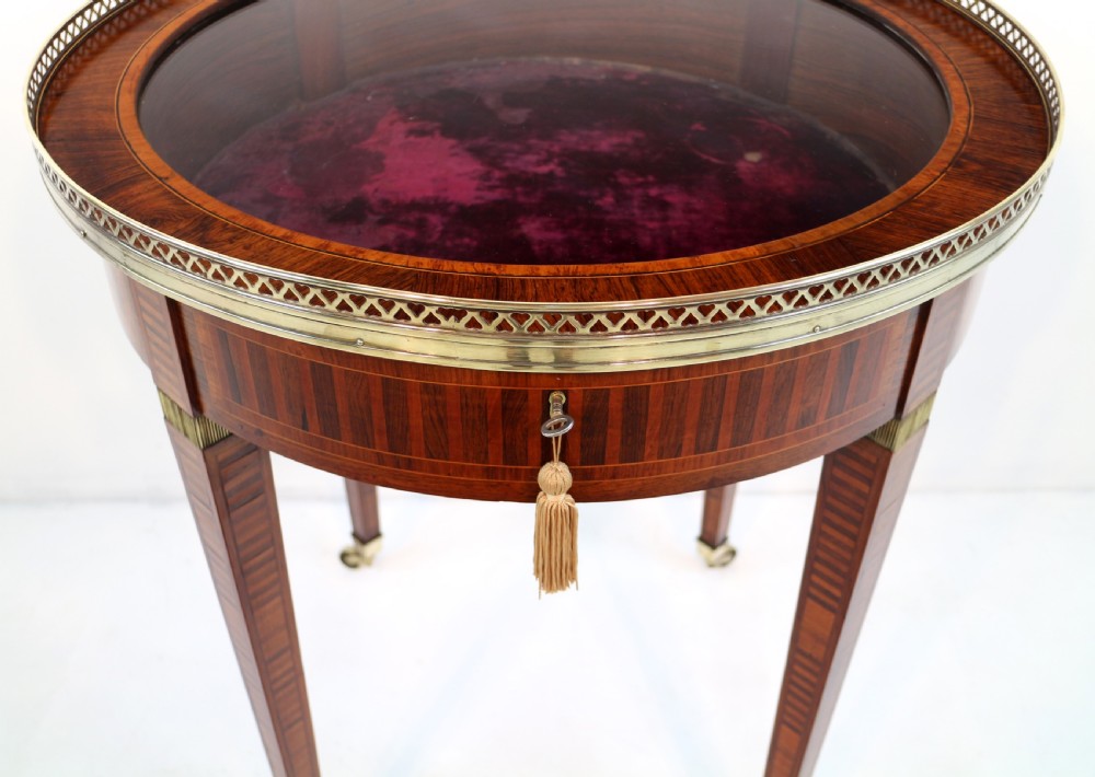 A circular Victorian rosewood bijouterie table featuring a glazed display lid, pierced brass rim gallery, inlaid tapered legs, and brass castors. View VIII