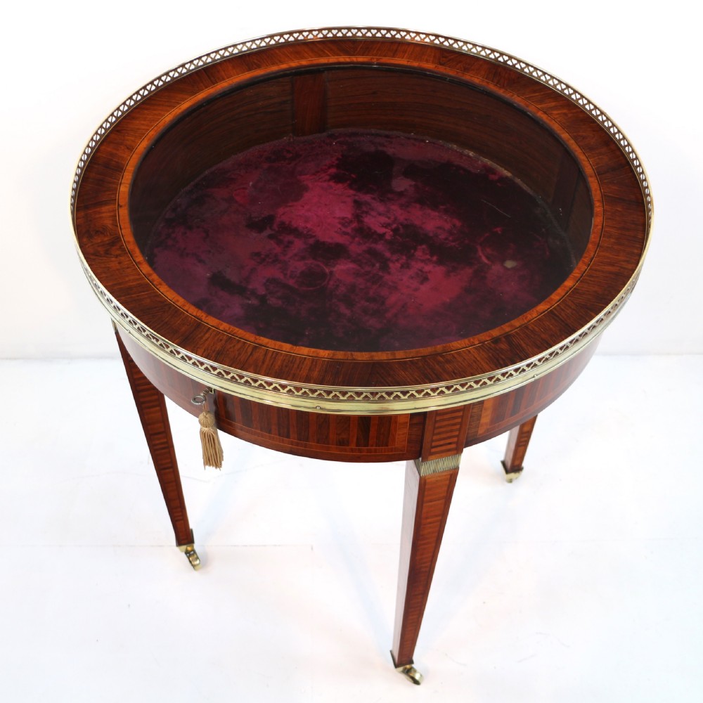 A circular Victorian rosewood bijouterie table featuring a glazed display lid, pierced brass rim gallery, inlaid tapered legs, and brass castors. View XI