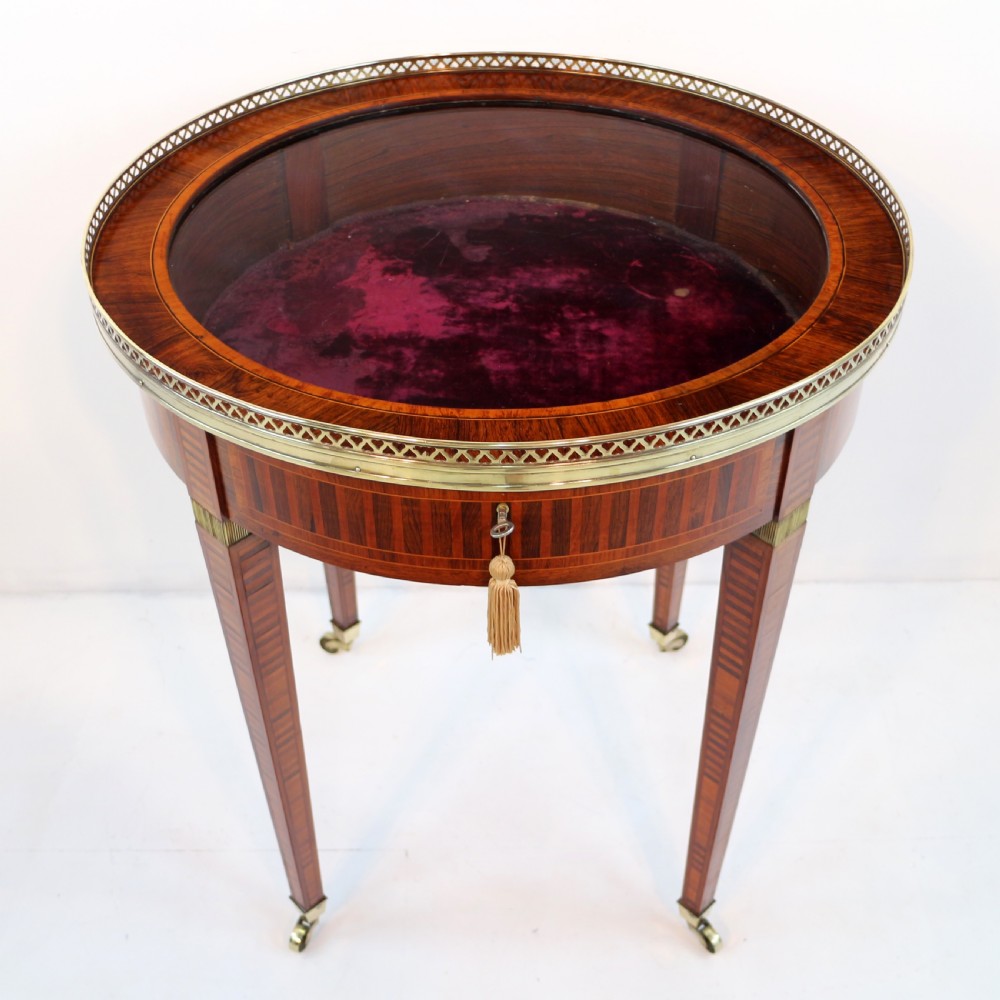 A circular Victorian rosewood bijouterie table featuring a glazed display lid, pierced brass rim gallery, inlaid tapered legs, and brass castors. View XII