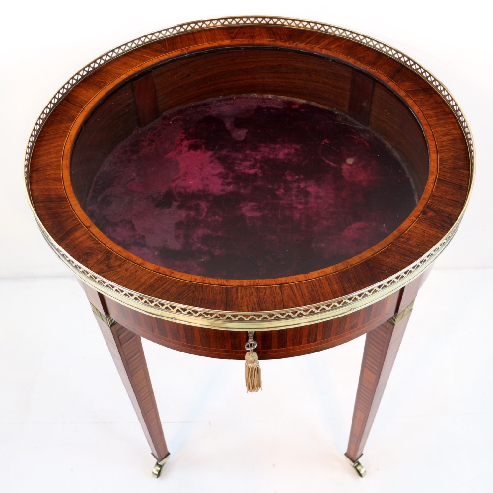 A circular Victorian rosewood bijouterie table featuring a glazed display lid, pierced brass rim gallery, inlaid tapered legs, and brass castors. View XIII