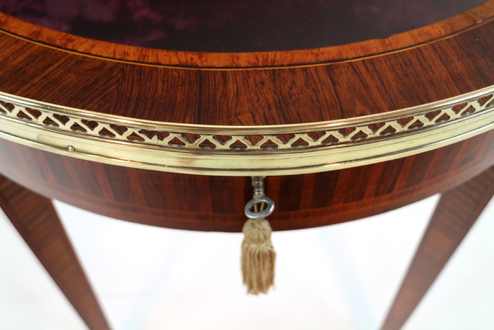 A circular Victorian rosewood bijouterie table featuring a glazed display lid, pierced brass rim gallery, inlaid tapered legs, and brass castors. View XIV