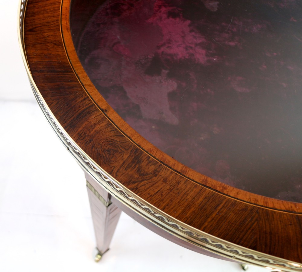 A circular Victorian rosewood bijouterie table featuring a glazed display lid, pierced brass rim gallery, inlaid tapered legs, and brass castors. View XV