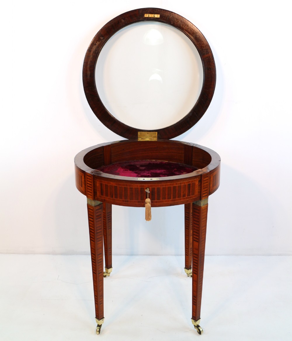 A circular Victorian rosewood bijouterie table featuring a glazed display lid, pierced brass rim gallery, inlaid tapered legs, and brass castors. View XVII