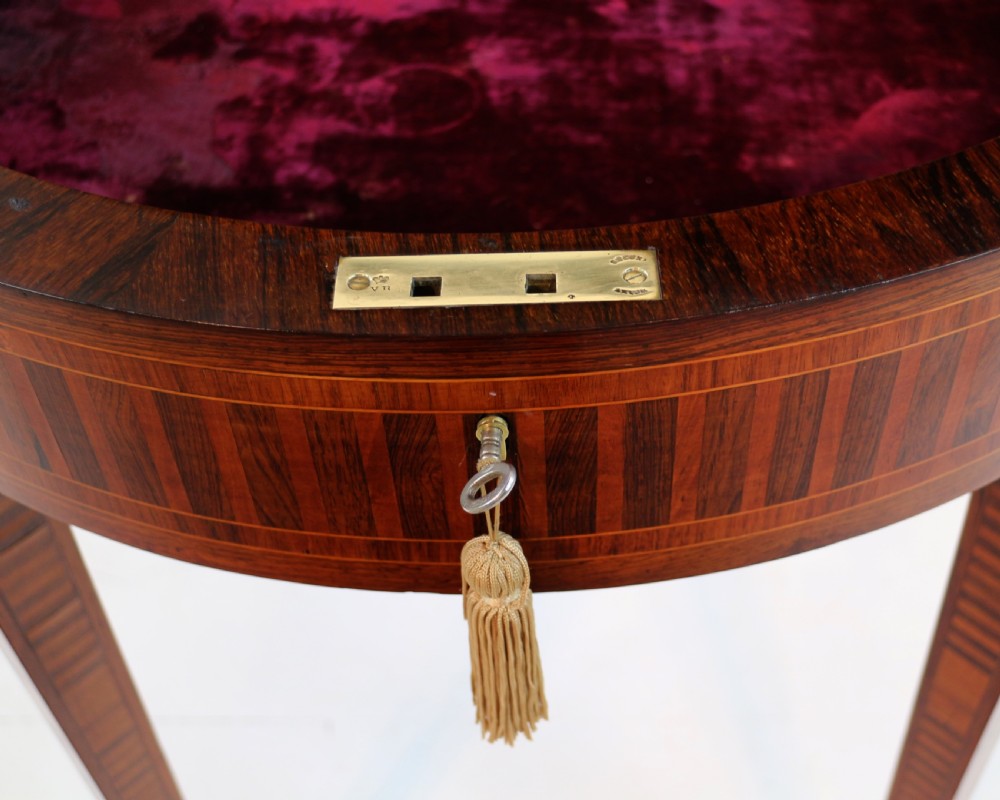 A circular Victorian rosewood bijouterie table featuring a glazed display lid, pierced brass rim gallery, inlaid tapered legs, and brass castors. View XIX
