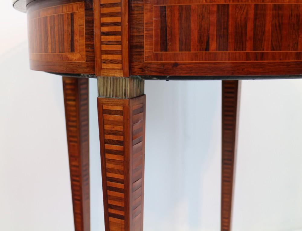 A circular Victorian rosewood bijouterie table featuring a glazed display lid, pierced brass rim gallery, inlaid tapered legs, and brass castors. View XXII