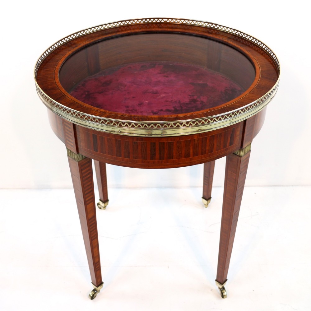A circular Victorian rosewood bijouterie table featuring a glazed display lid, pierced brass rim gallery, inlaid tapered legs, and brass castors. View XXV