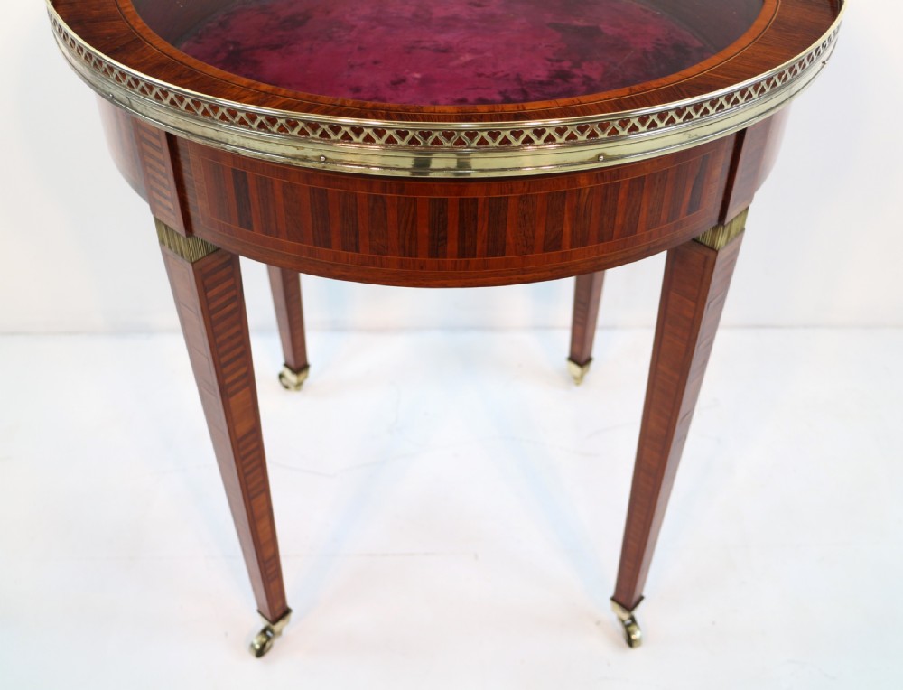 A circular Victorian rosewood bijouterie table featuring a glazed display lid, pierced brass rim gallery, inlaid tapered legs, and brass castors. View XXVI