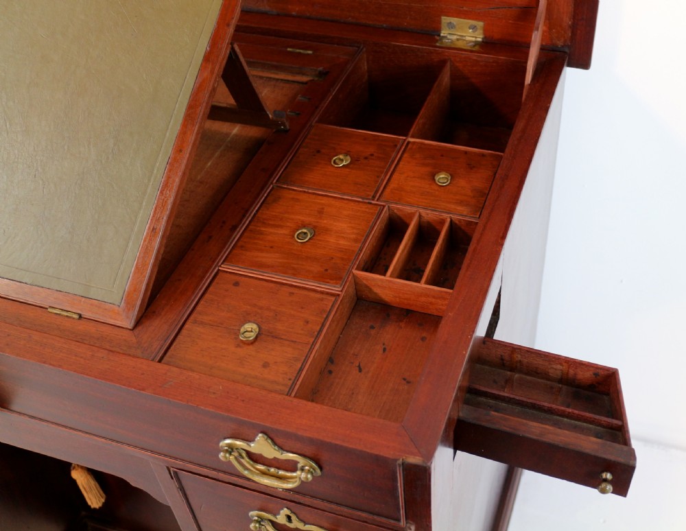 An antique George II red walnut kneehole desk featuring a hinged top that opens to reveal internal compartments and an adjustable green leather mirror, resting on bracket feet with brass handles. View XX