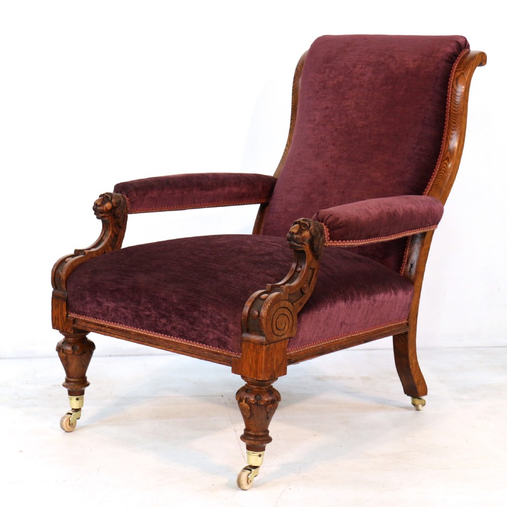 A pair of Victorian oak library chairs upholstered in plum velvet featuring carved lion head arm terminals and brass castors. View XIV