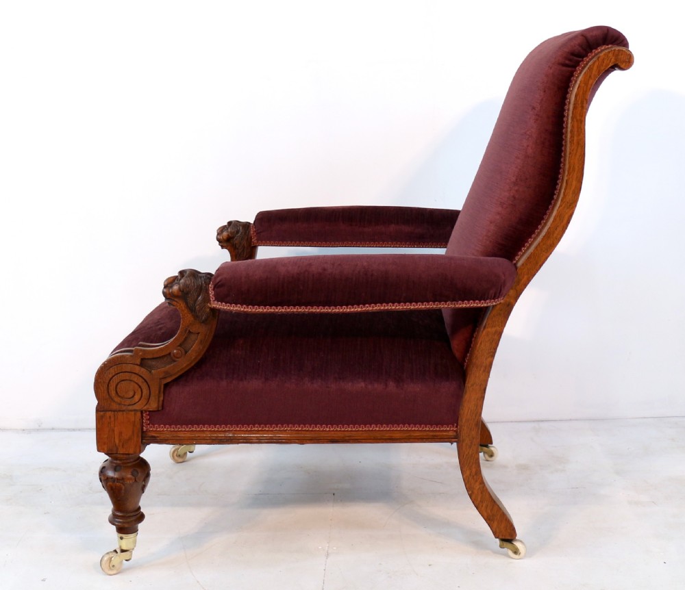 A pair of Victorian oak library chairs upholstered in plum velvet featuring carved lion head arm terminals and brass castors. View XV