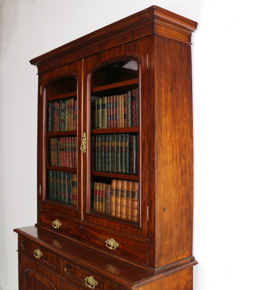 An early Victorian mahogany bookcase featuring an upper glazed section with two arched doors and a lower cupboard base with two drawers. View XXX