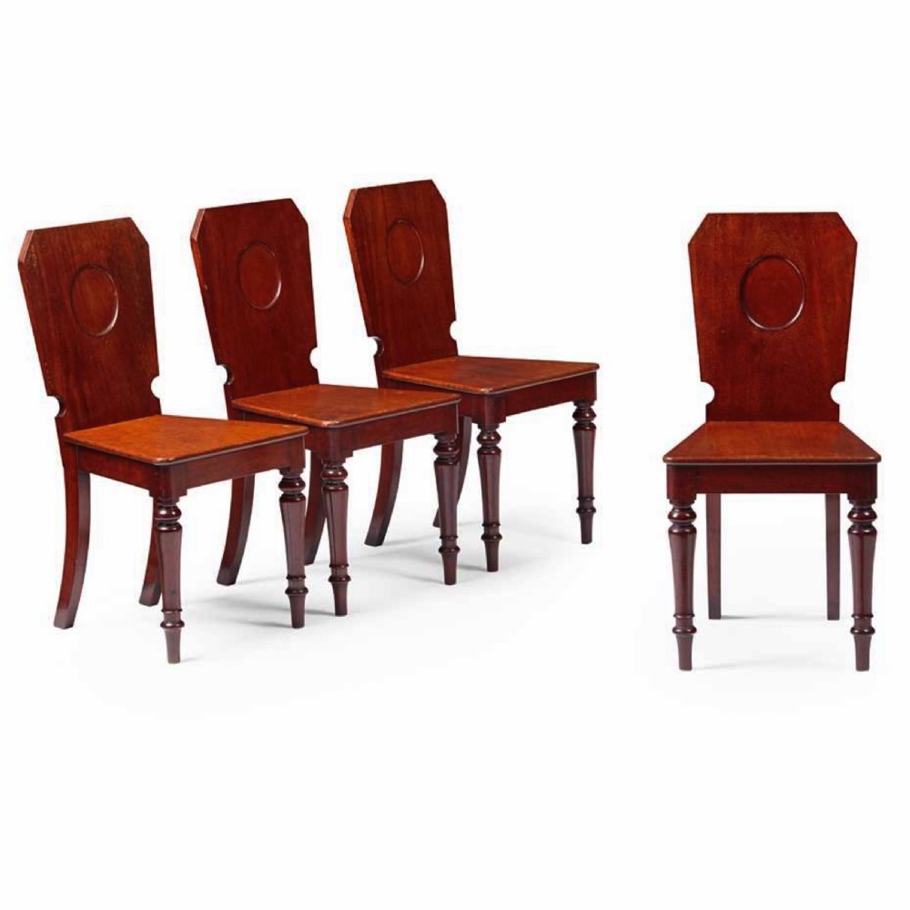 A set of four Victorian mahogany hall chairs featuring shaped solid wooden backs with recessed circular central details, flat wooden seats, turned front legs, and flared rear legs.