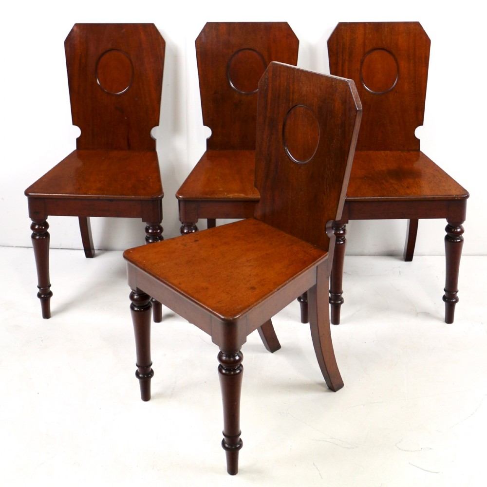 A set of four Victorian mahogany hall chairs featuring shaped solid wooden backs with recessed circular central details, flat wooden seats, turned front legs, and flared rear legs. View II